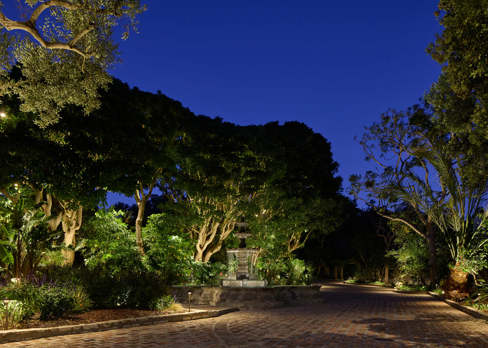 A brick motor court at night, its fountain and old trees softly uplit