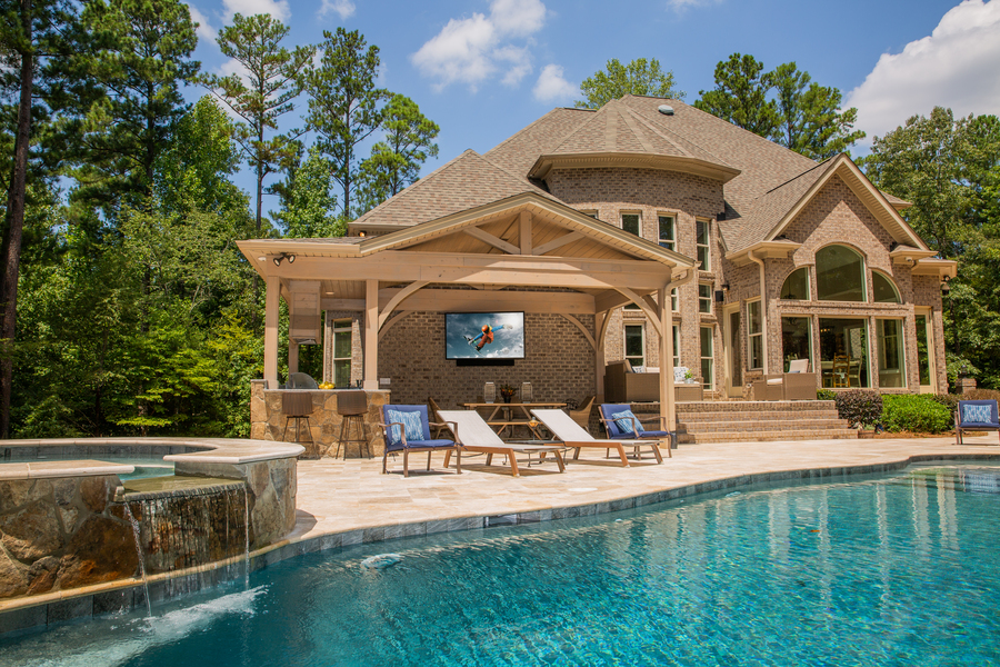 A home with a large pool in the front has a patio on the side. There’s an outdoor TV mounted in the shade of the patio.